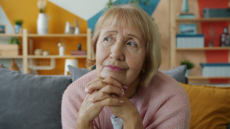 Elderly woman with hands clasped, looking thoughtful.